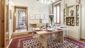Un coin repas avec une table en bois dressée pour quatre personnes, des chaises transparentes, un lustre et un tapis rouge à motifs.