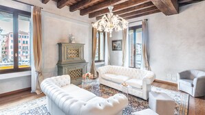Un salon spacieux avec un plafond haut, un lustre, un canapé blanc, un fauteuil beige et un tapis à motifs.