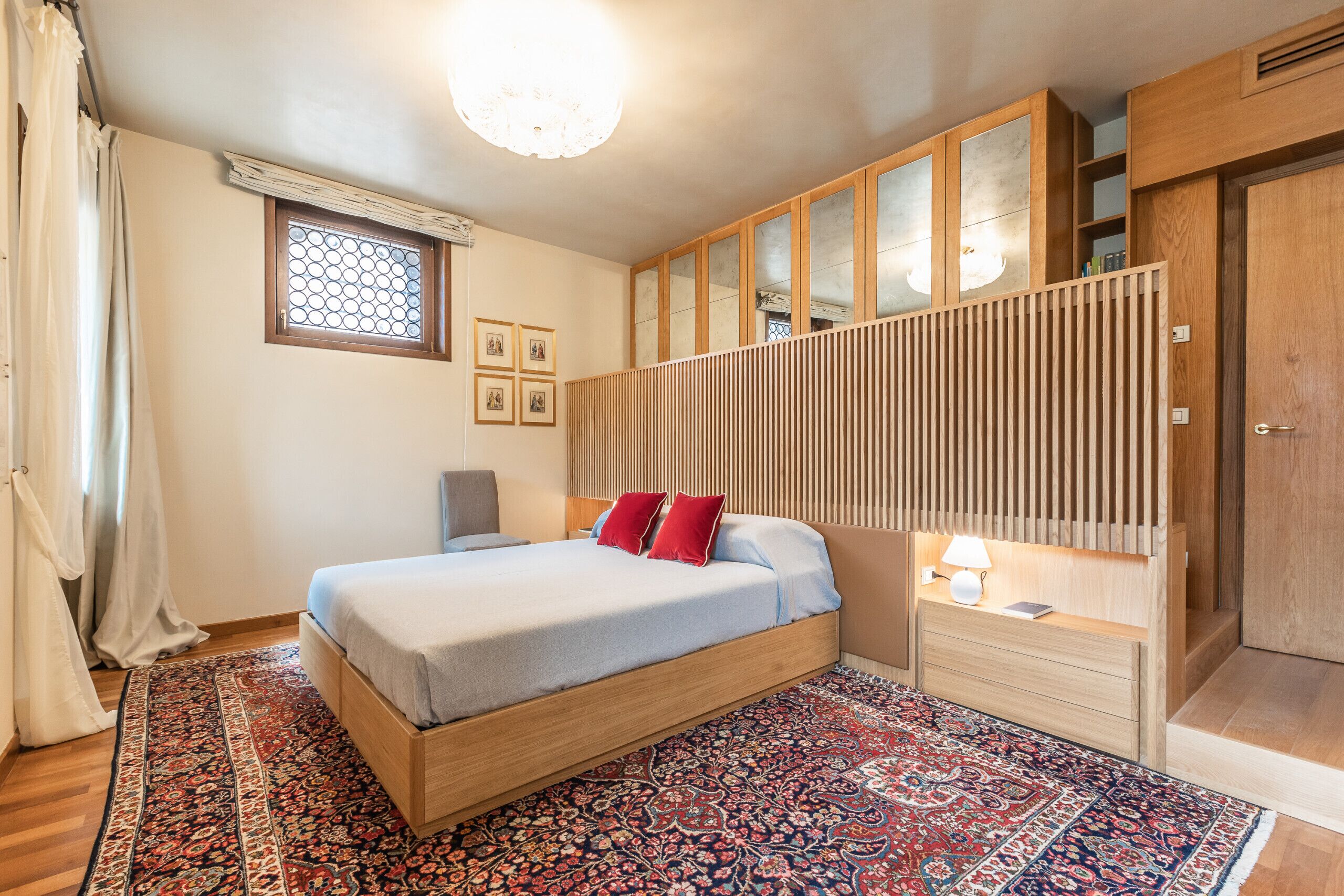 A bedroom with a bed, a nightstand, a lamp, a window with curtains, and a wooden wardrobe.
