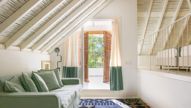 Une pièce avec un plafond haut, un canapé vert, un tapis à motifs et, vue depuis l’encadrement d’une porte, un balcon extérieur.