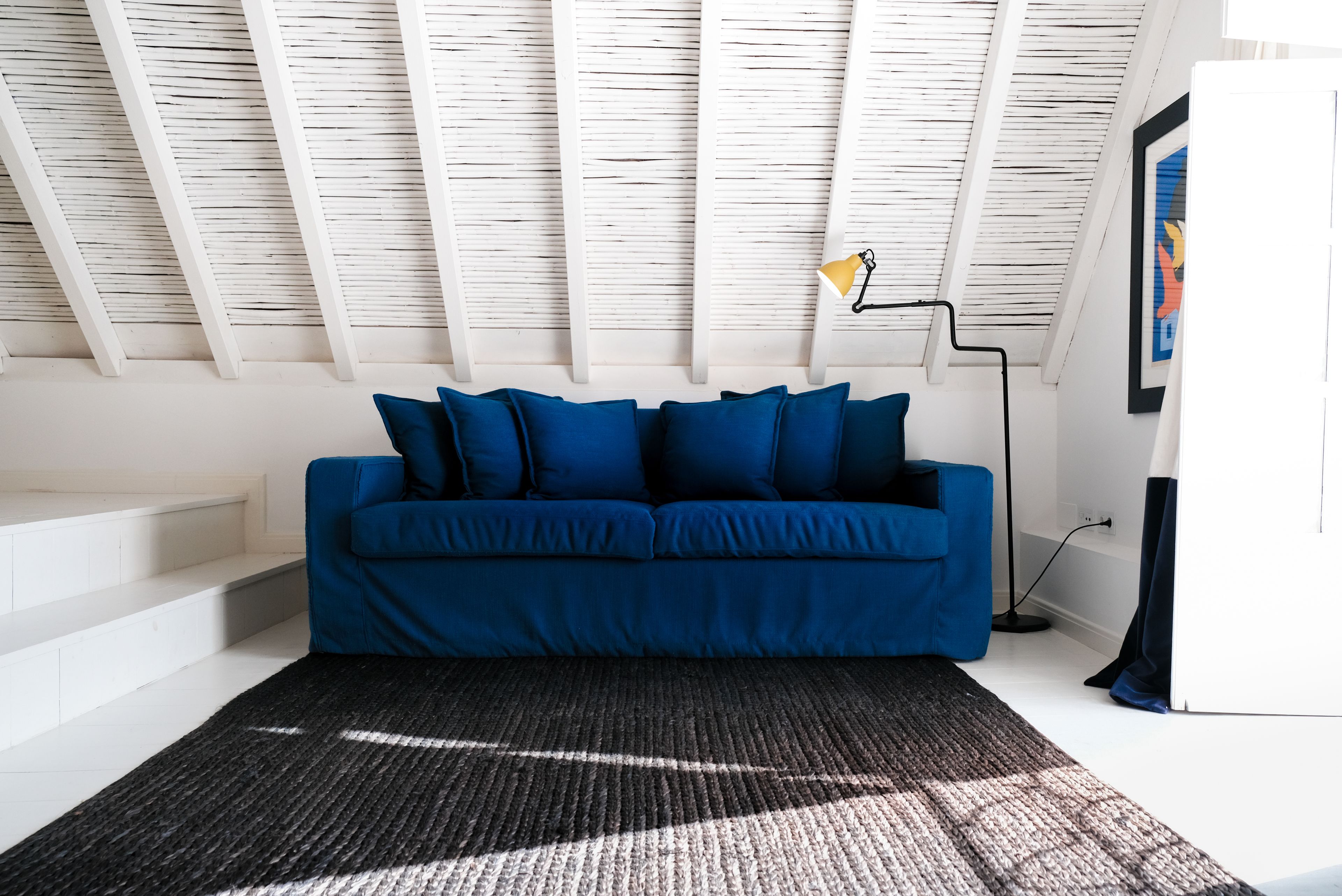 A modern living room with a blue sofa, a black and gray rug, and a white ceiling with wooden beams.