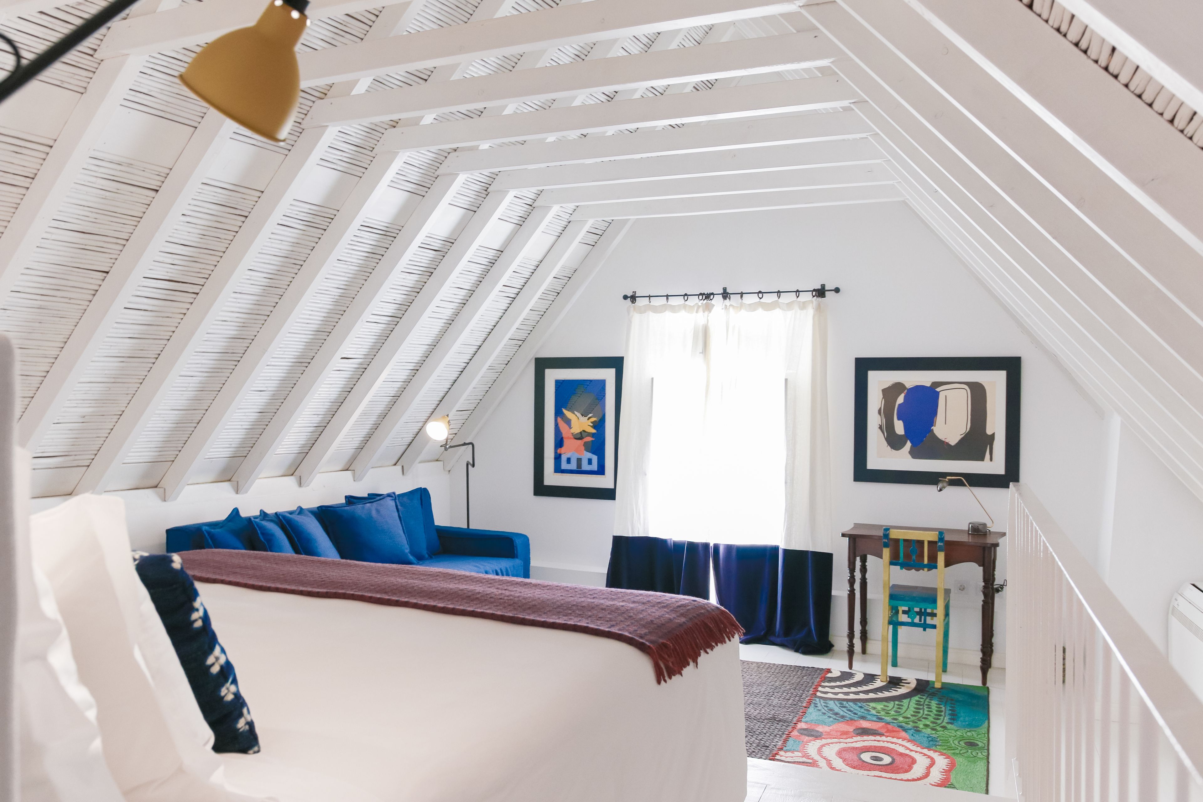A room with a sloped ceiling, a bed with blue bedding, a small table, and a chair.