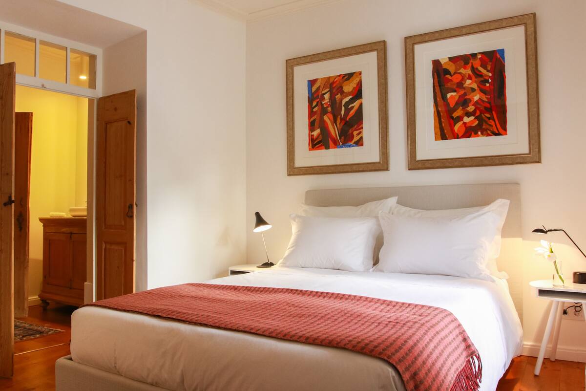 Ein Schlafzimmer mit einem großen Bett, einem Nachttisch mit Lampe sowie zwei gerahmten abstrakten Bildern an der Wand.