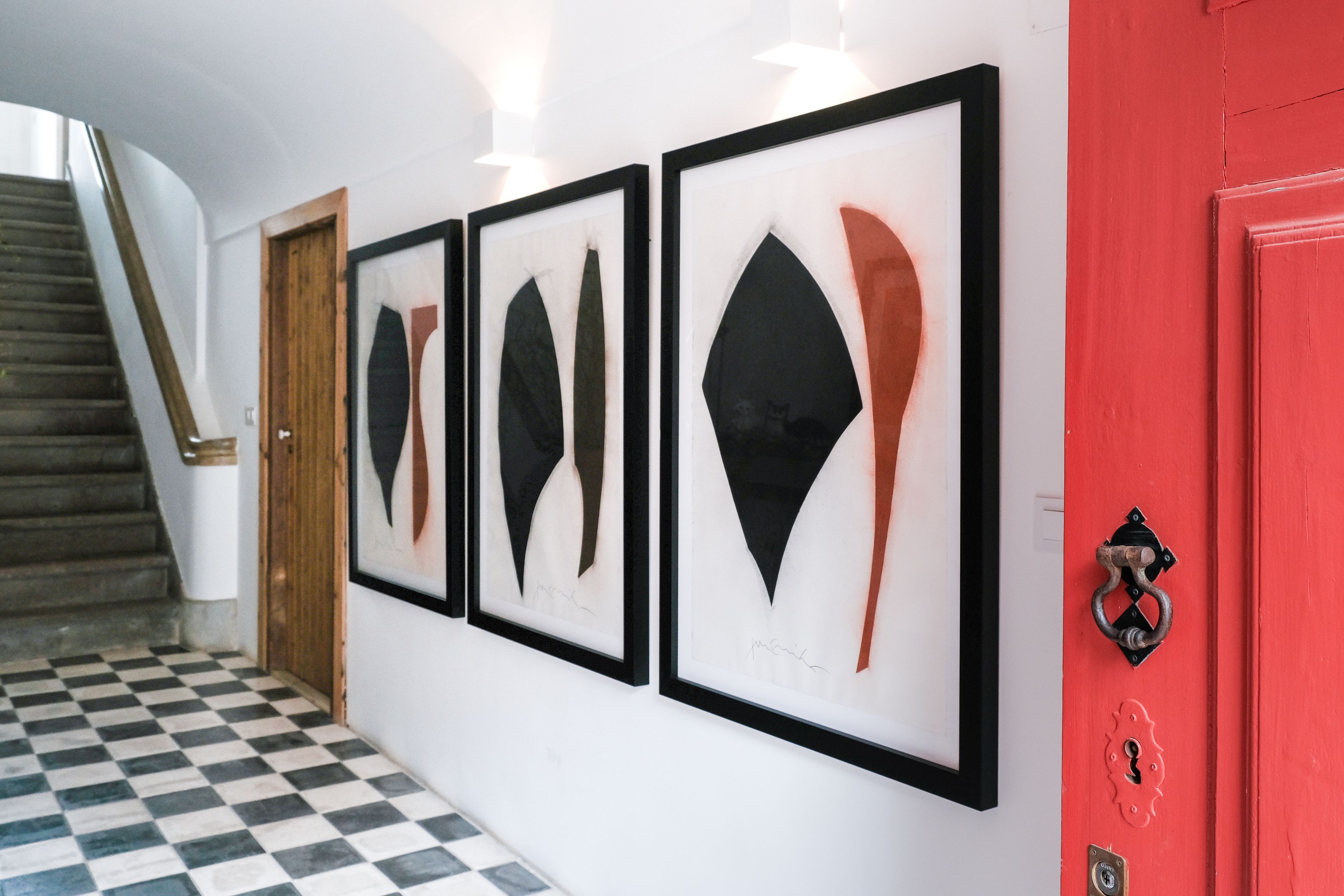 A hallway with framed artwork on the wall, a staircase, and a red door with a keyhole.