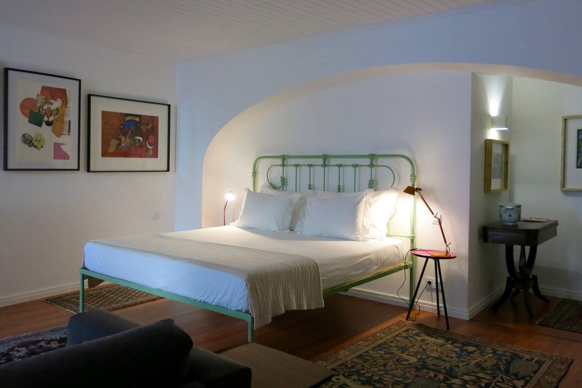 Ein Schlafzimmer mit einem grünem Metallbettgestell, weißer Bettwäsche, einem Nachttisch mit Lampe und zwei gerahmten Bildern an der Wand.