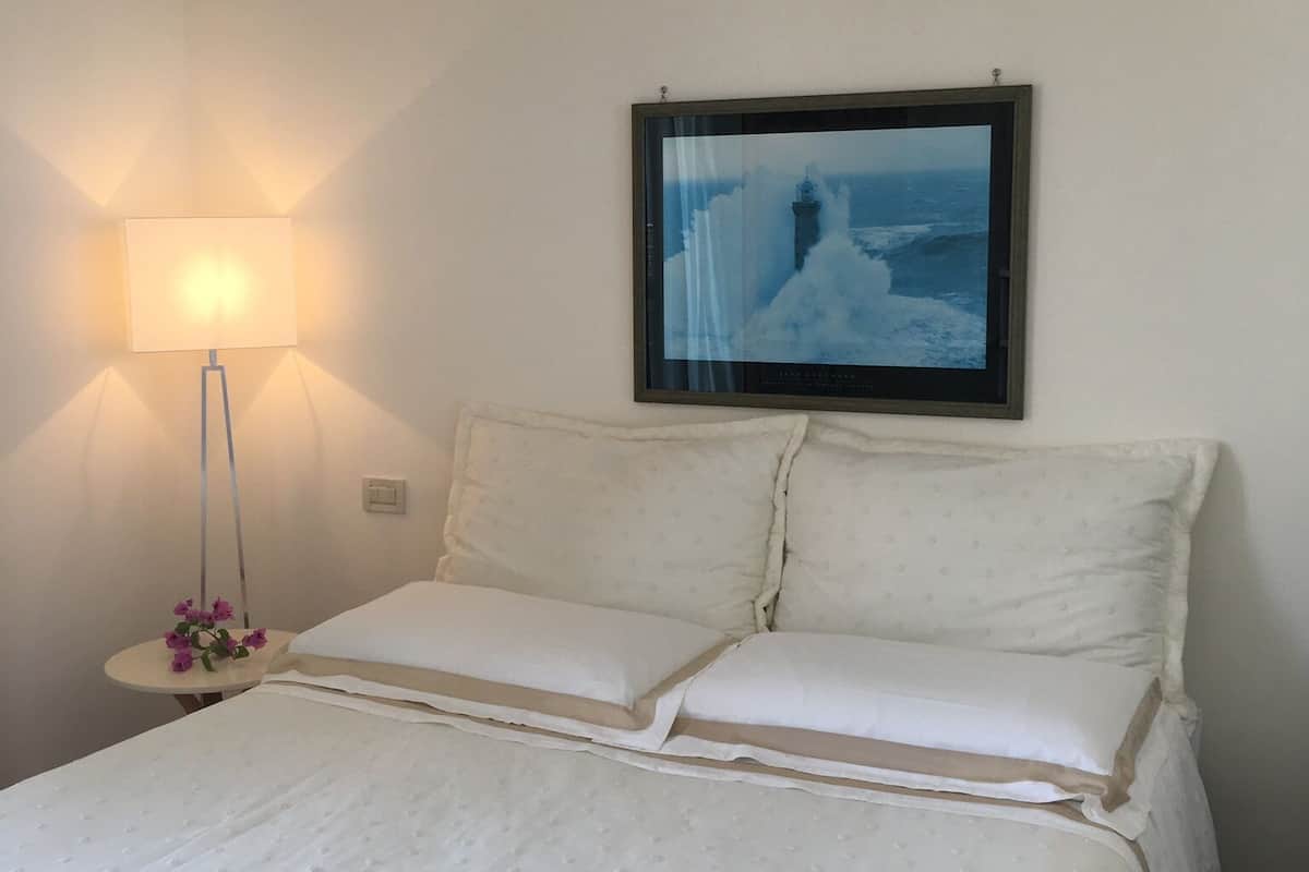 Ein ordentlich bezogenes Bett mit weißen Leinen und Kissen, ein gerahmtes Bild mit Küstenmotiv an der Wand und eine Nachttischlampe mit Blumenarrangement.
