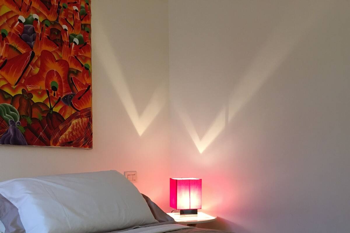 Ein ordentlich bezogenes Bett mit einem weißen Kopfkissen und einer braunen Decke, eine pinkfarbene Lampe auf einem Nachttisch und ein farbenfrohes abstraktes Gemälde an der Wand.