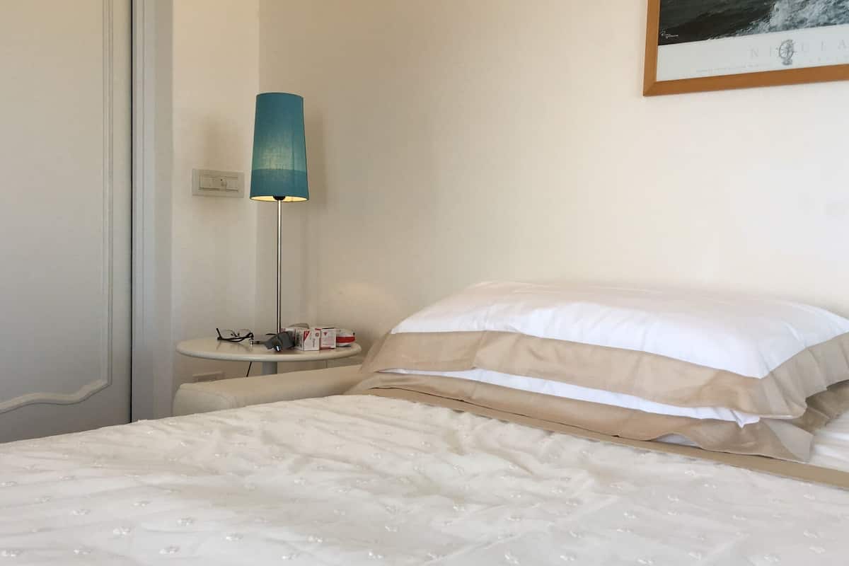 Ein ordentlich bezogenes Bett mit weißer Bettwäsche, ein Nachttisch mit Lampe und ein gerahmtes Bild an der Wand.