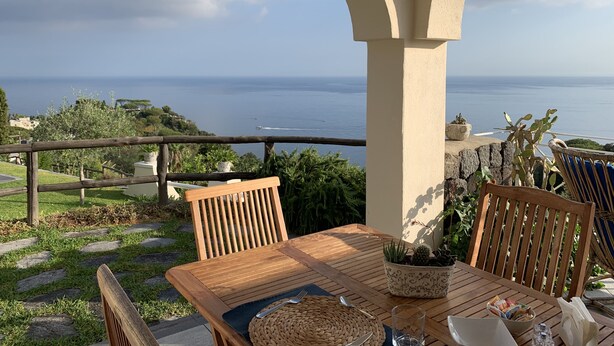 Une table en bois avec des chaises, un set de table en fibres tressées, un verre et un bol rempli de magazines, le tout sur un balcon donnant sur la mer.
