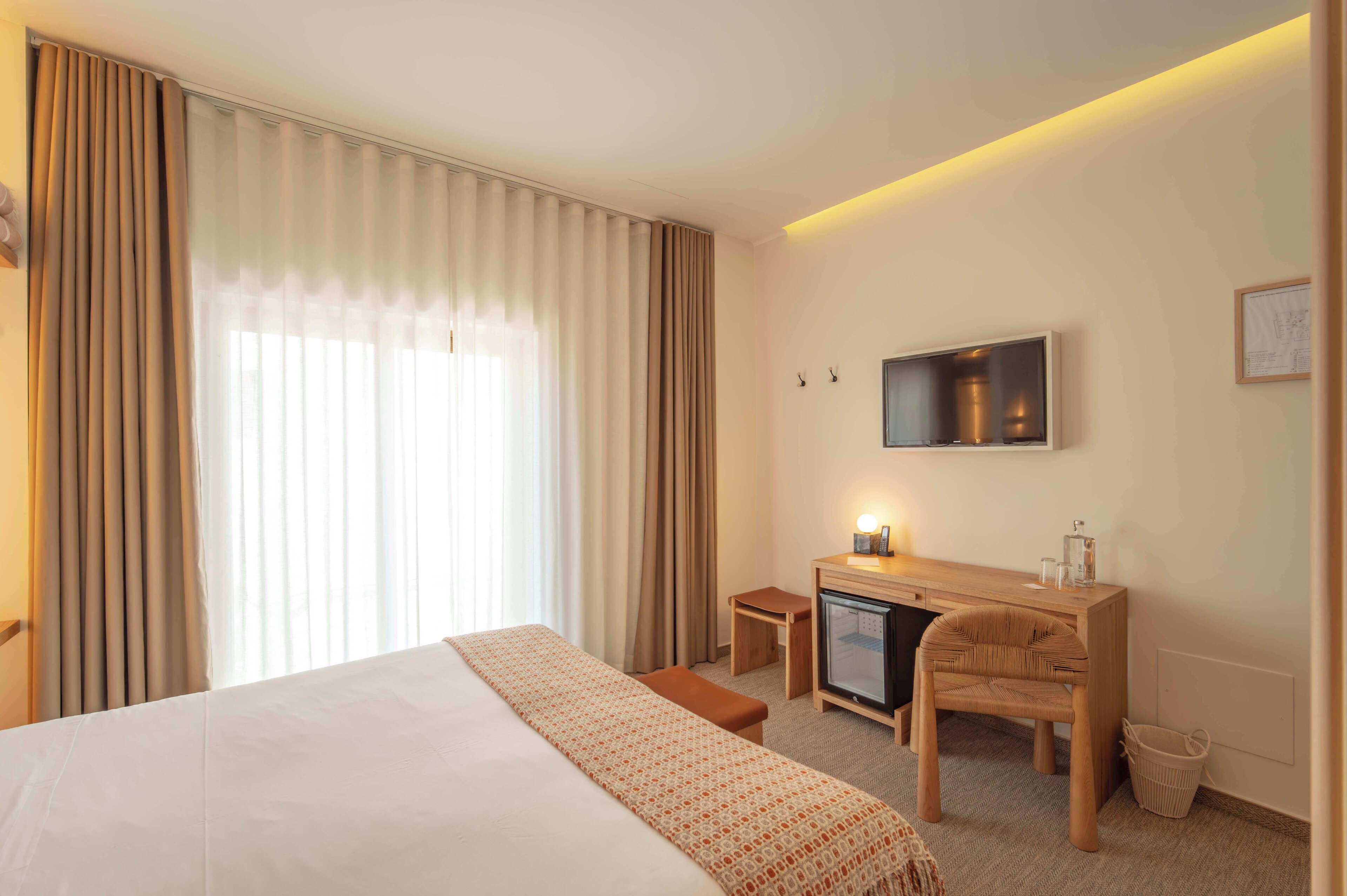 A hotel room with a bed, a desk with a chair, a television, and a window with curtains.