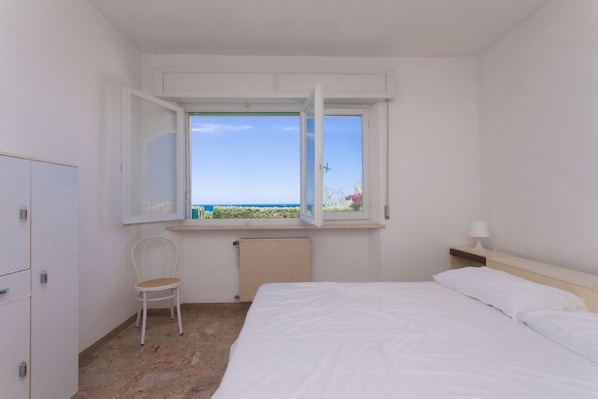 A bedroom with a bed, a chair, a nightstand, a window with an open view, and a radiator.