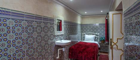 Une pièce dont les murs sont recouverts de carreaux à motifs, avec un lavabo blanc, deux lits avec une literie rouge et une porte dotée d’un cadre doré.