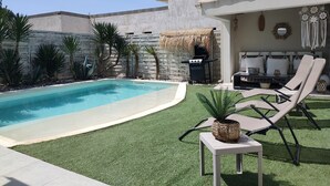 Ein Pool mit Grill unter einem Reetdach, Liegestühlen und einem kleinen Tisch mit einer Pflanze.