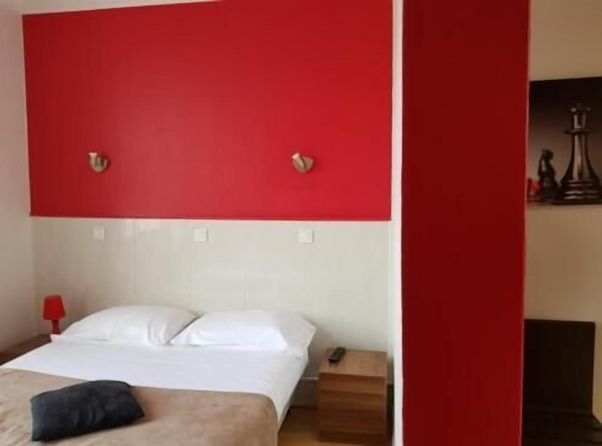 Ein Hotelzimmer mit roter Akzentwand, einem Bett mit weißer Bettwäsche, einem Nachttisch mit Lampe und einem kleinen Holz-Nachttisch.