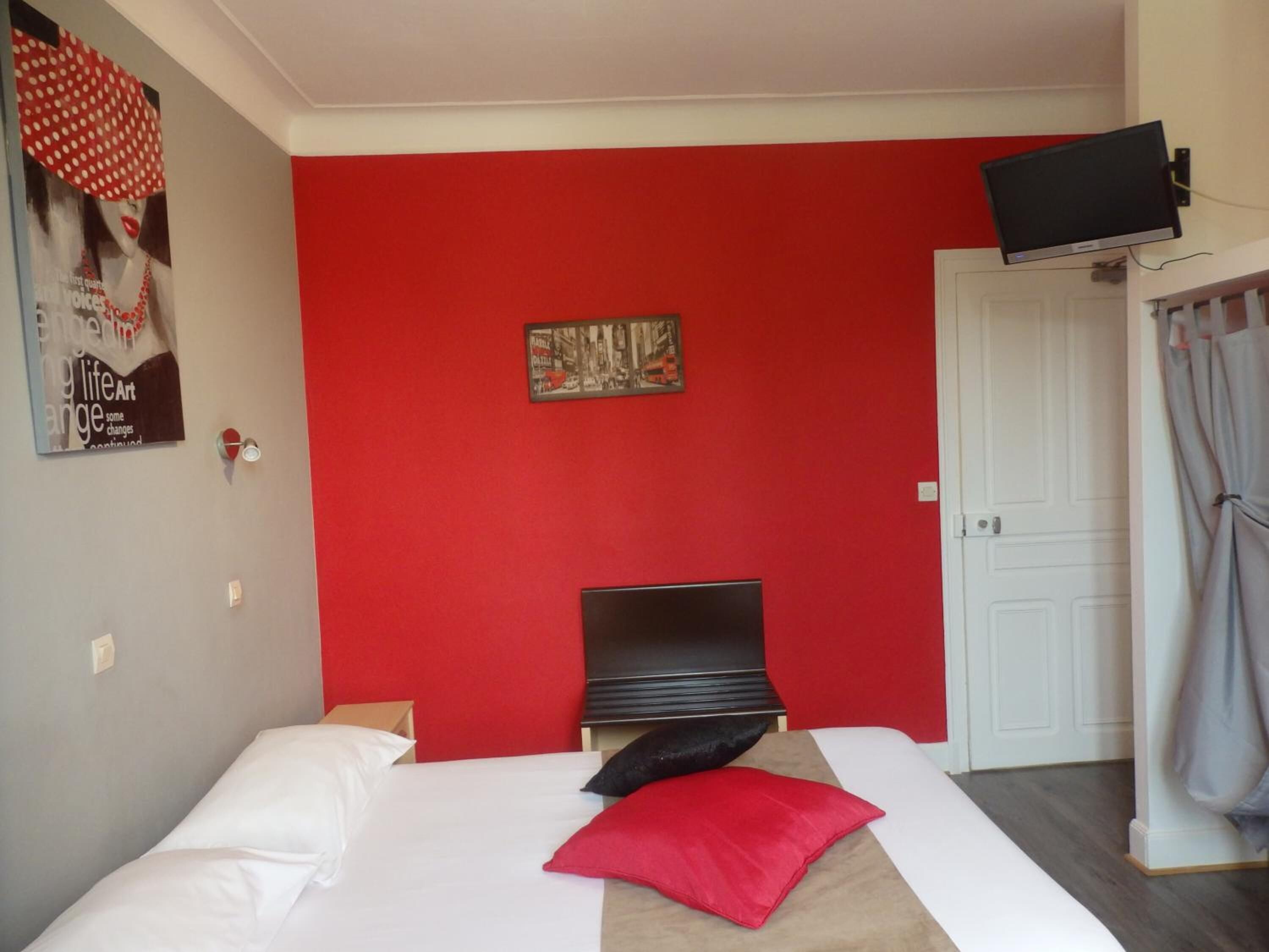 Une chambre avec un mur rouge, un lit avec une literie blanche et rouge, un téléviseur à écran plat fixé au mur et une porte avec des rideaux.