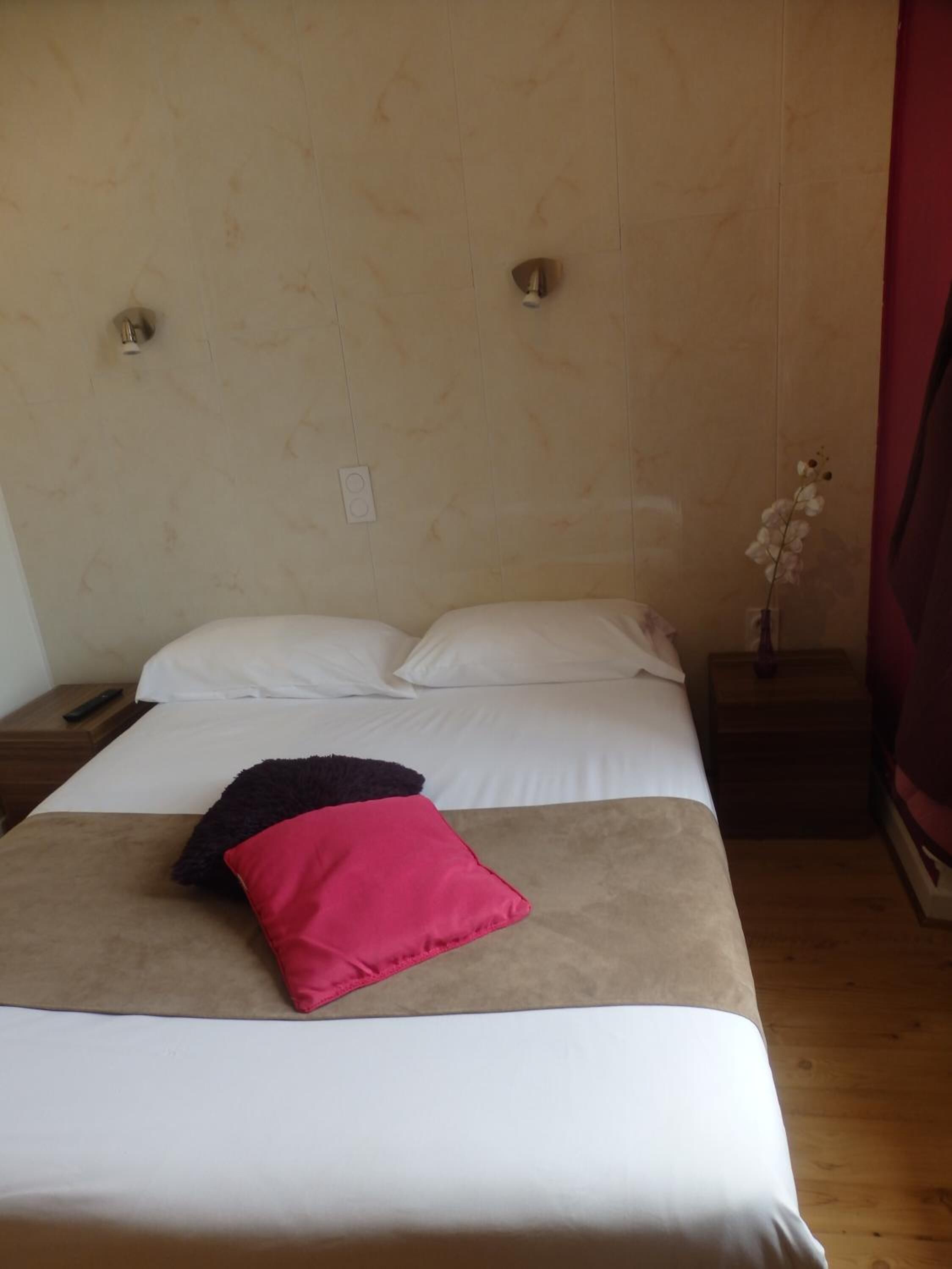 Una cama bien hecha con sábanas blancas y una sola almohada rosa, un control remoto en una mesita de noche de madera, y un jarrón con una orquídea sobre la mesita.