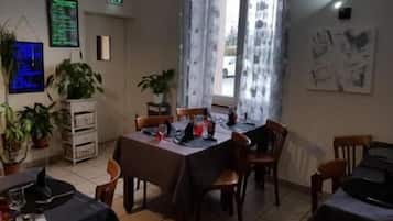 Un comedor con una mesa dispuesta para dos comensales, sillas, plantas en macetas y una ventana con cortinas.
