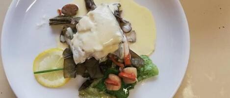 A white plate with a fish dish, lemon slice, and greens.