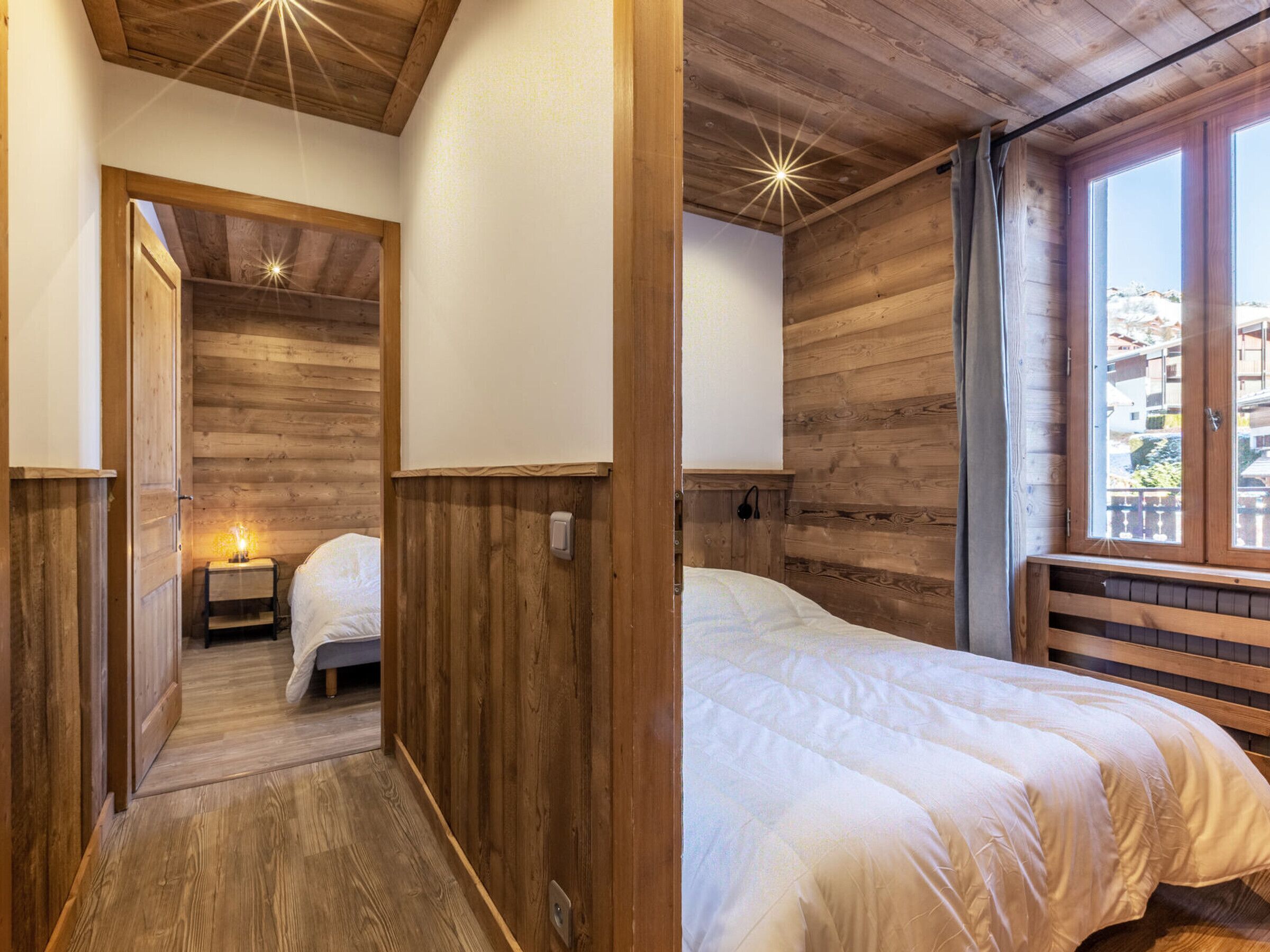 A wooden room with a bed, a bedside table with a lamp, and a window with a view of buildings.