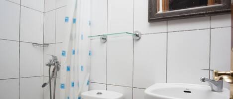 A bathroom with white tiles, a shower, a toilet, and a sink with a gold faucet.