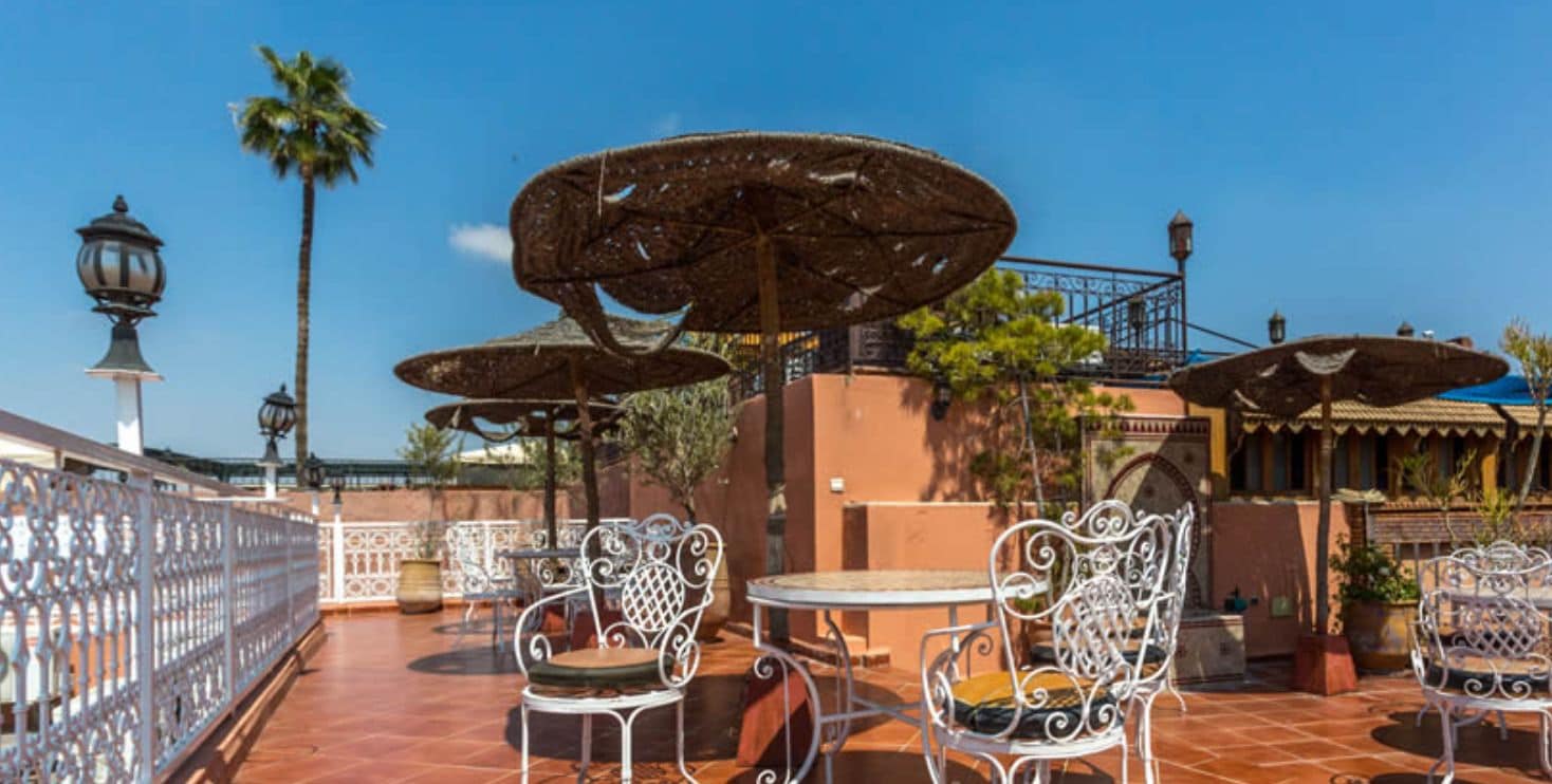 Une terrasse extérieure avec des chaises et des tables en métal, protégée par des parasols en osier, et agrémentée d’une clôture décorative.