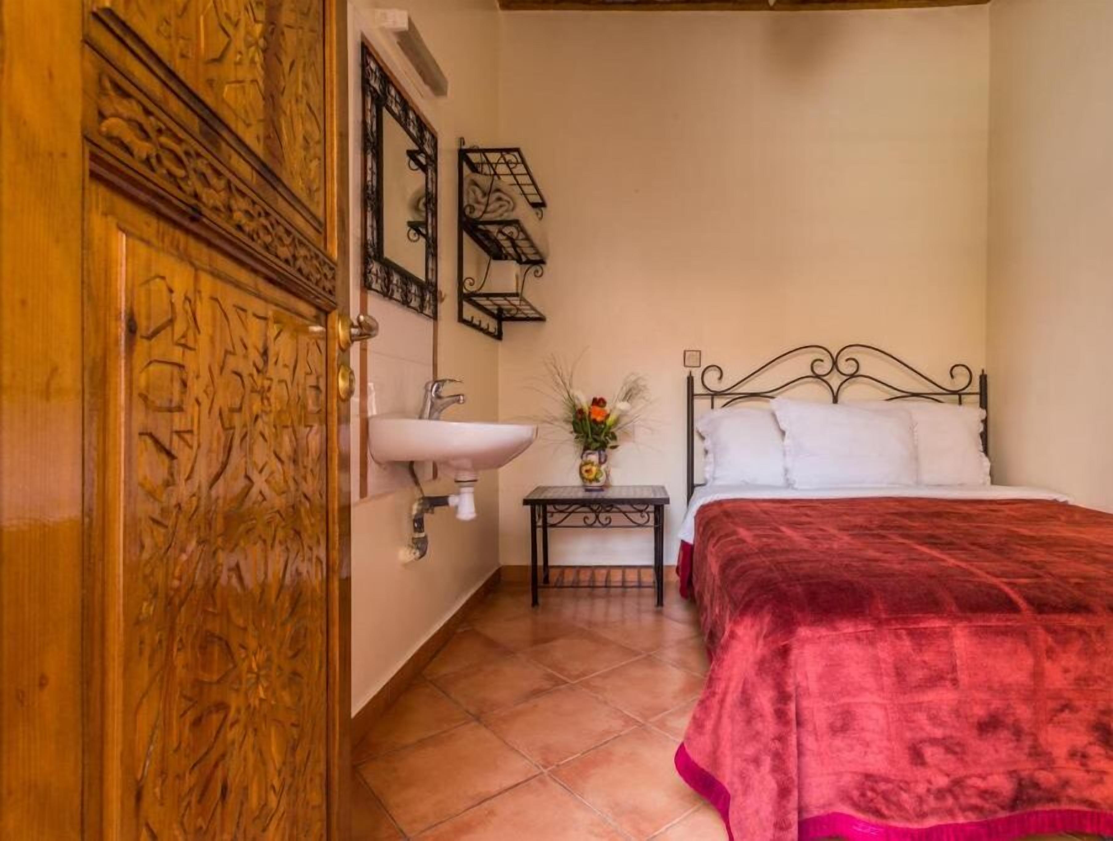 A bedroom with a wooden wardrobe, a white sink, a bedside table with a vase, and a bed with a red blanket.