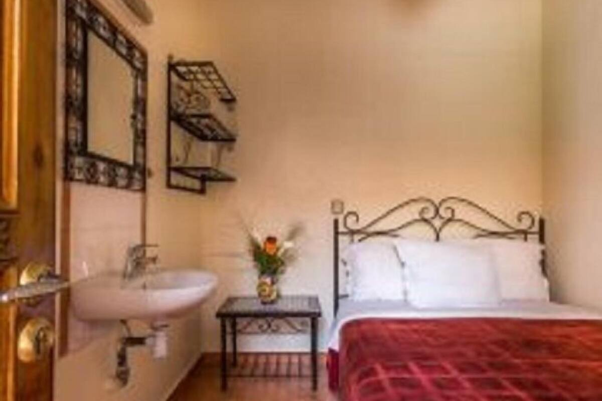 A small, cozy bedroom with a single bed, a bedside table with a vase of flowers, a wall-mounted shelf, and a bathroom with a sink and mirror.