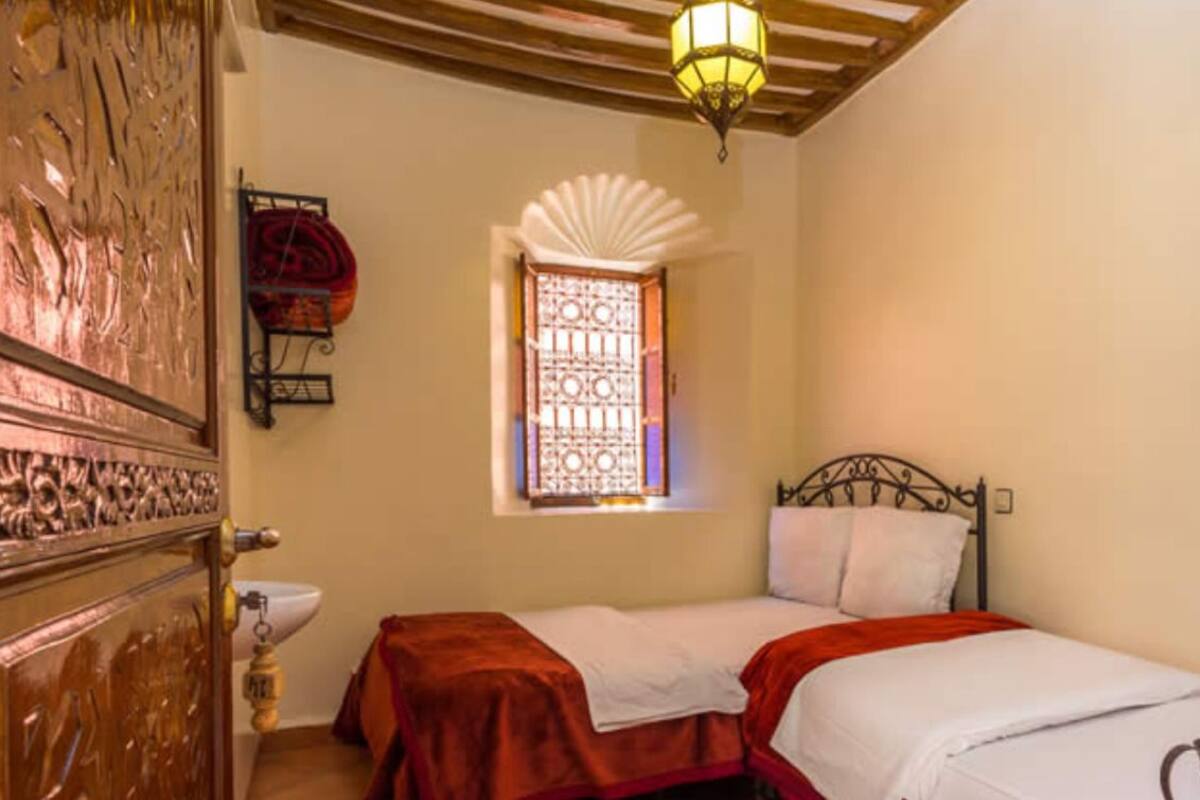 A room with a bed, a window with decorative bars, a wall-mounted shelf with a red item, and a wooden ceiling.