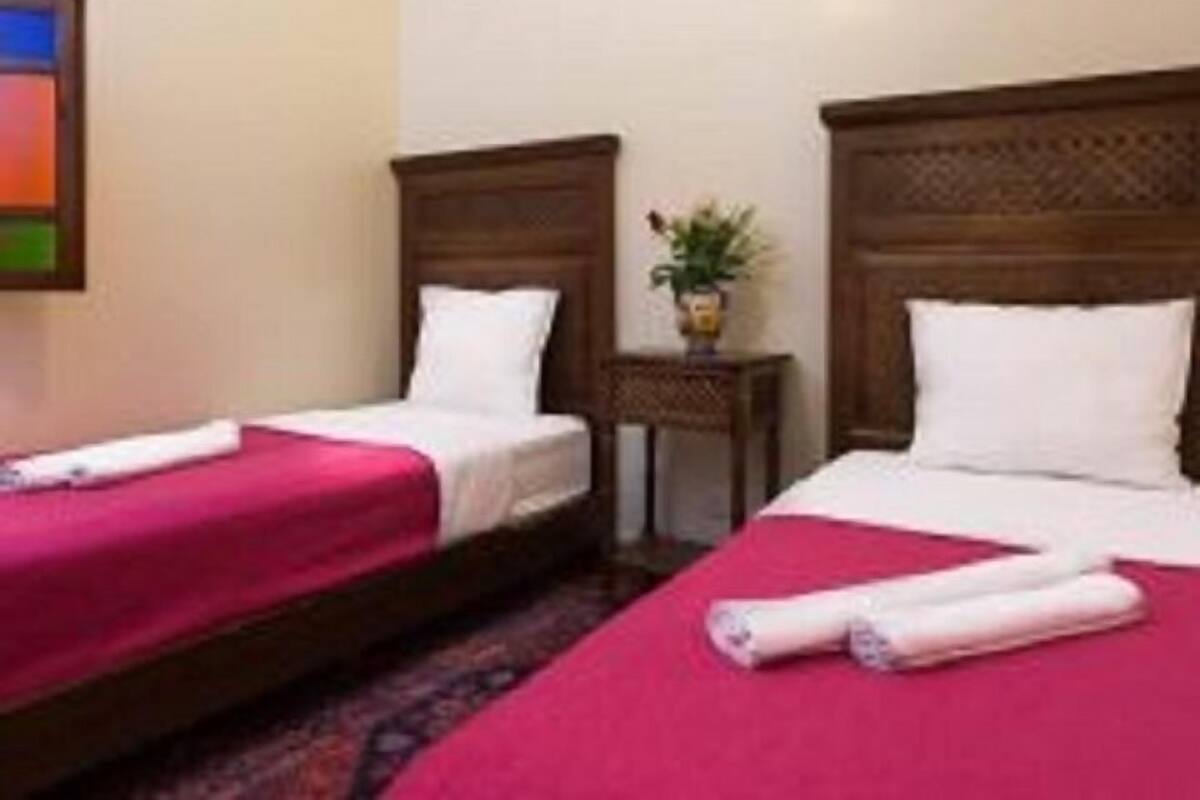 A hotel room with two single beds, each with a wooden headboard, a small bedside table with a plant, and a red bedspread.