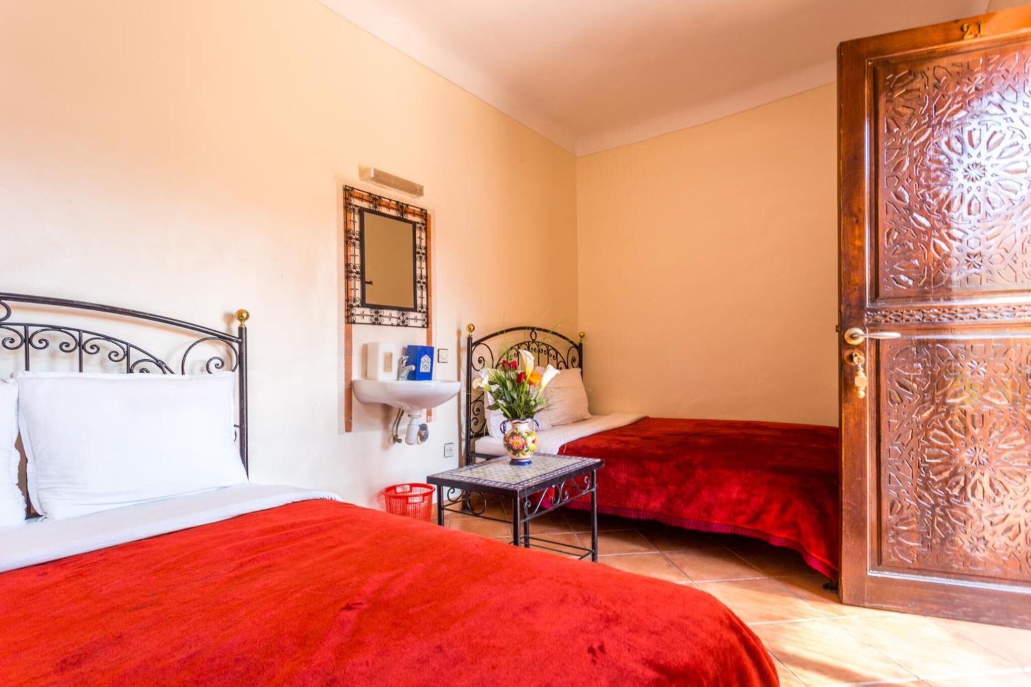 Une chambre d’hôtel avec deux lits, une petite table ornée d’un vase de fleurs, un miroir et une porte en bois aux sculptures complexes.