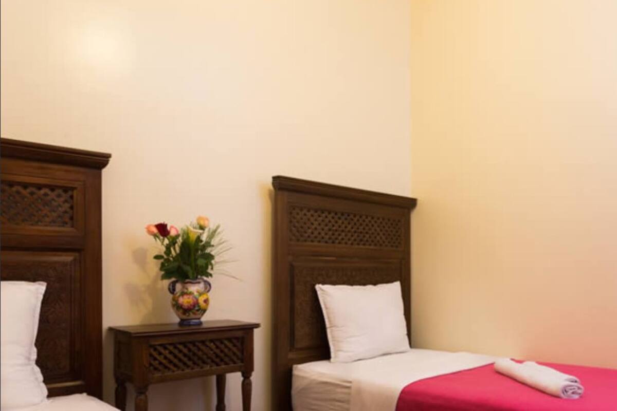 A hotel room with two beds, each with a wooden headboard, a small table with a vase of flowers, and a red bedspread.