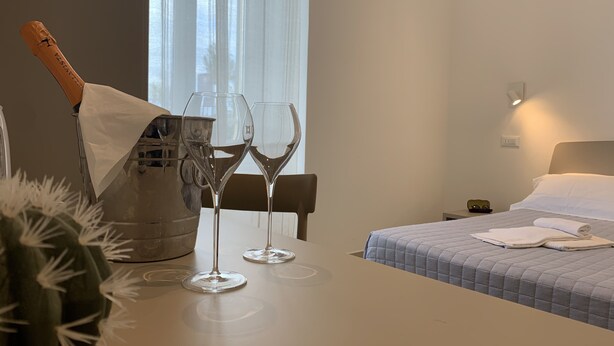 A hotel room with a bed, a bedside table with two wine glasses and a bottle of champagne, and a cactus plant.