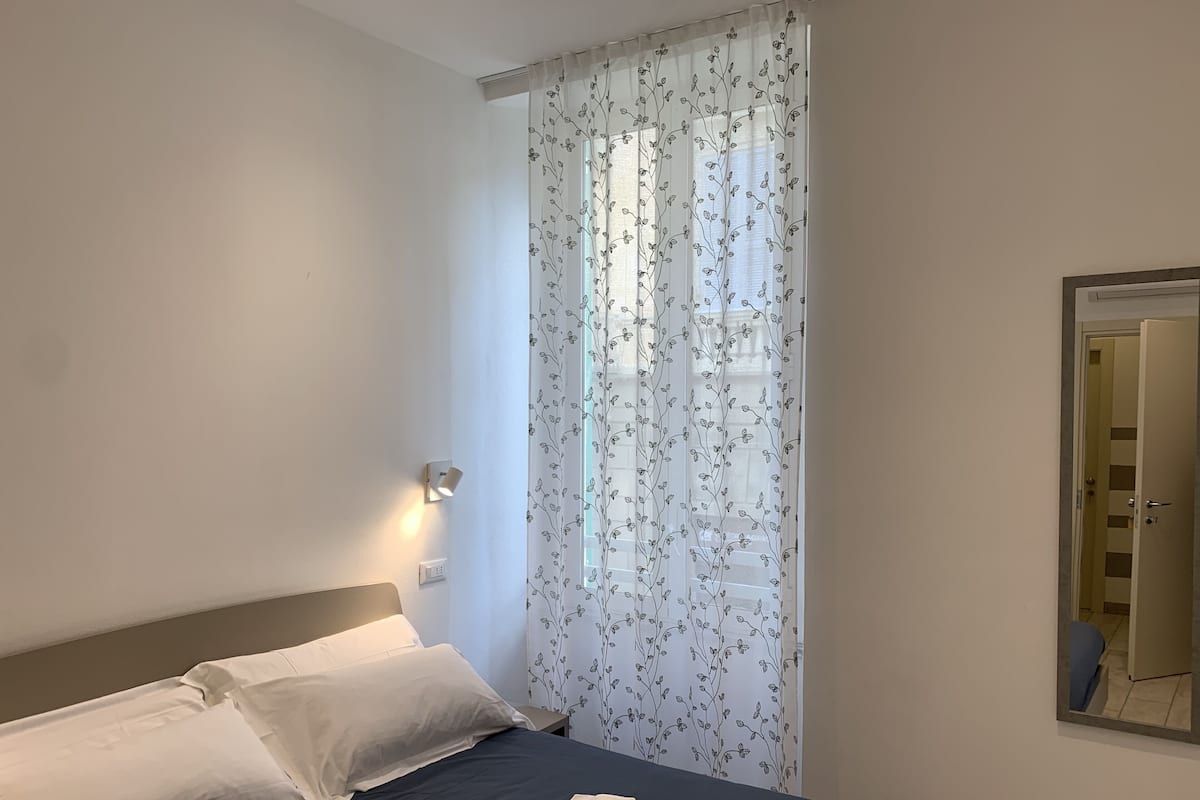 Un dormitorio con una cama, almohadas blancas, una toalla doblada, una lámpara de pared y una ventana con cortinas florales.
