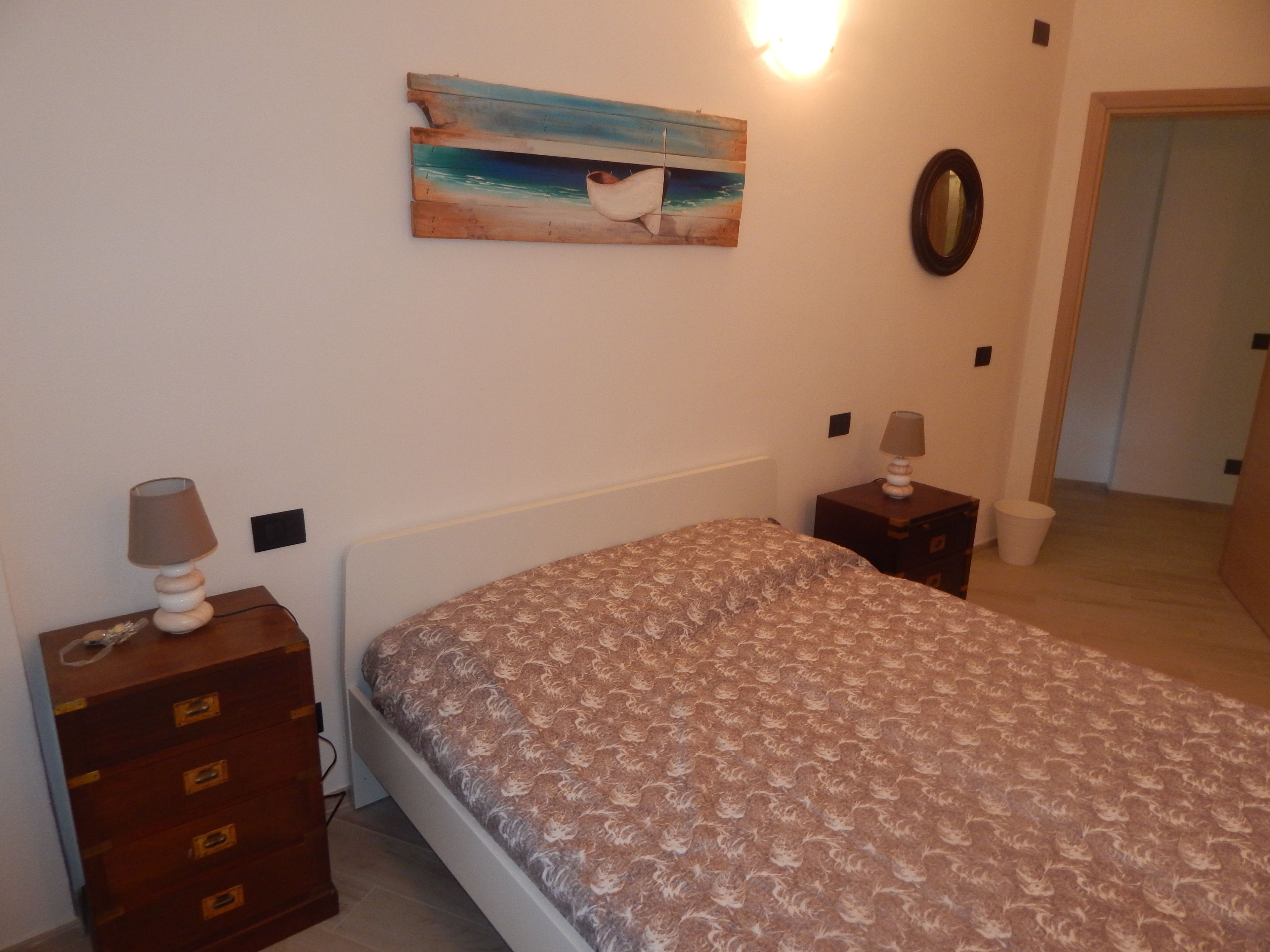 A bedroom with a bed, a dresser, a nightstand, a lamp, a painting of a beach scene, and a mirror.