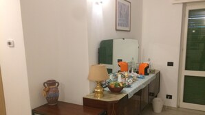 A small room with a wooden table, a lamp, a vase, a basket of fruit, and a refrigerator. - Guest House Berzefi (Bergeggi)