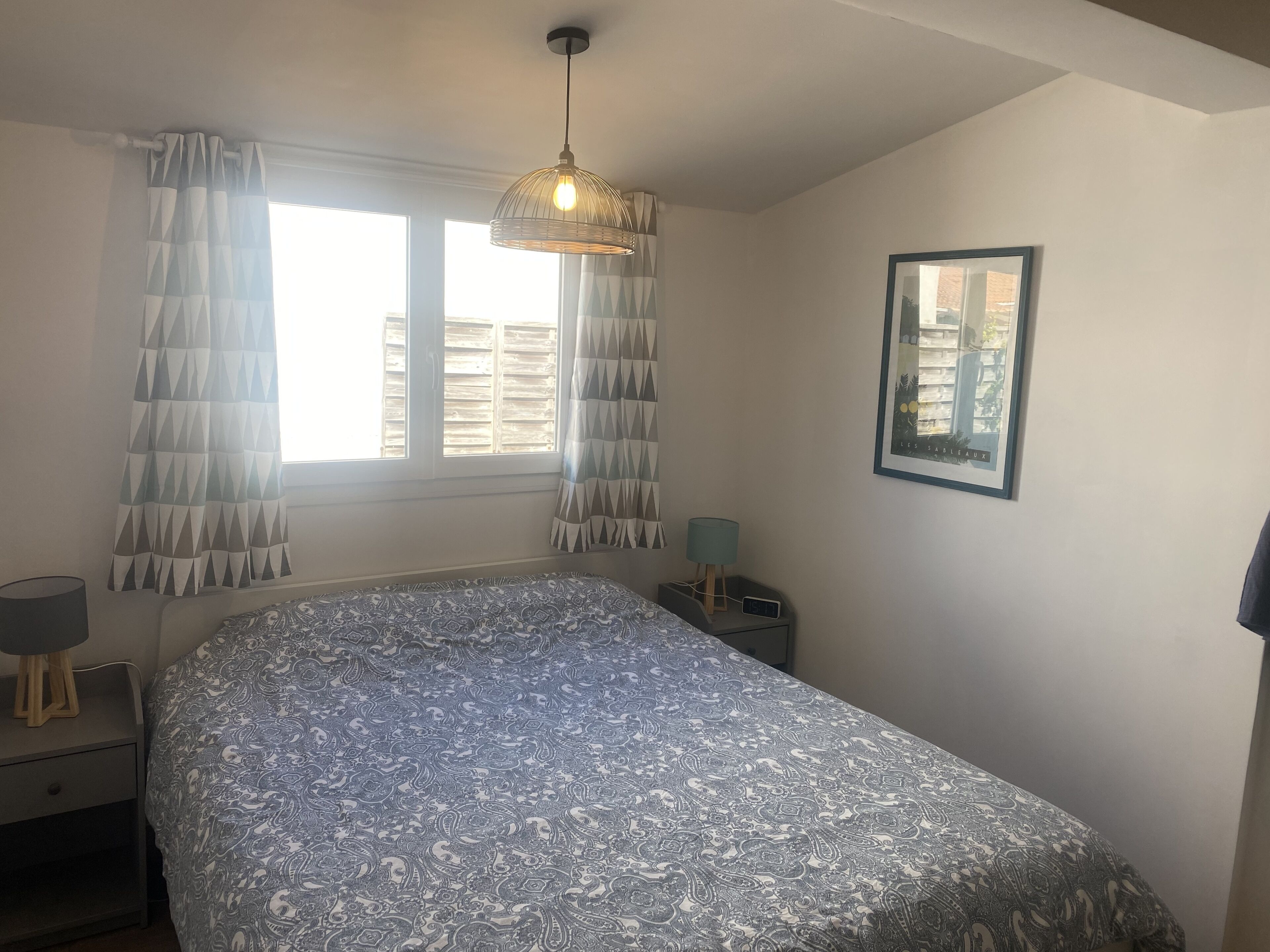 A bedroom with a bed, a window with curtains, a bedside lamp, a nightstand, and a framed picture on the wall.