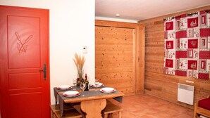 A dining area with a wooden table set for four, a red door with a floral design, and a wooden wall with a red and white patterned curtain. - 1st floor, balcony, parking, tv, ski locker, 23m², Pra Loup (Uvernet-Fours)