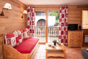 A wooden cabin with a sofa, a table with cups, a TV, and a balcony with curtains.