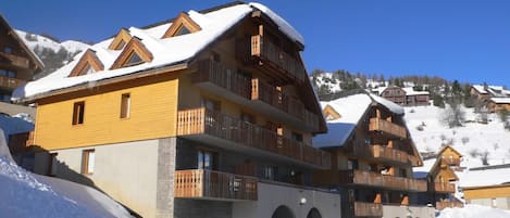 Un chalet à plusieurs étages, doté de balcons, qui se dresse sur fond de montagnes enneigées.