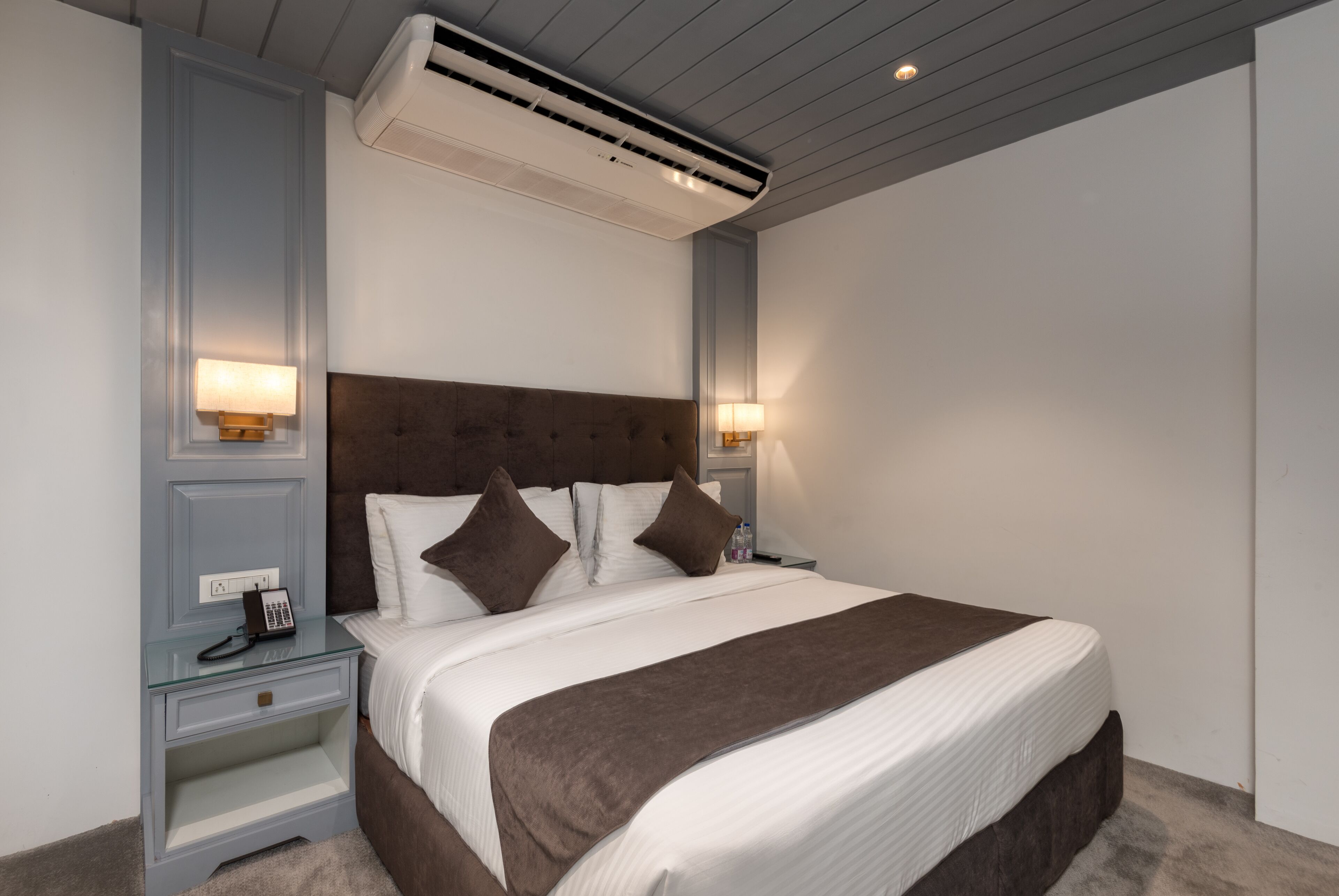 A neatly made bed with a brown headboard and white bedding, a bedside table with a lamp, and an air conditioning unit above the bed.