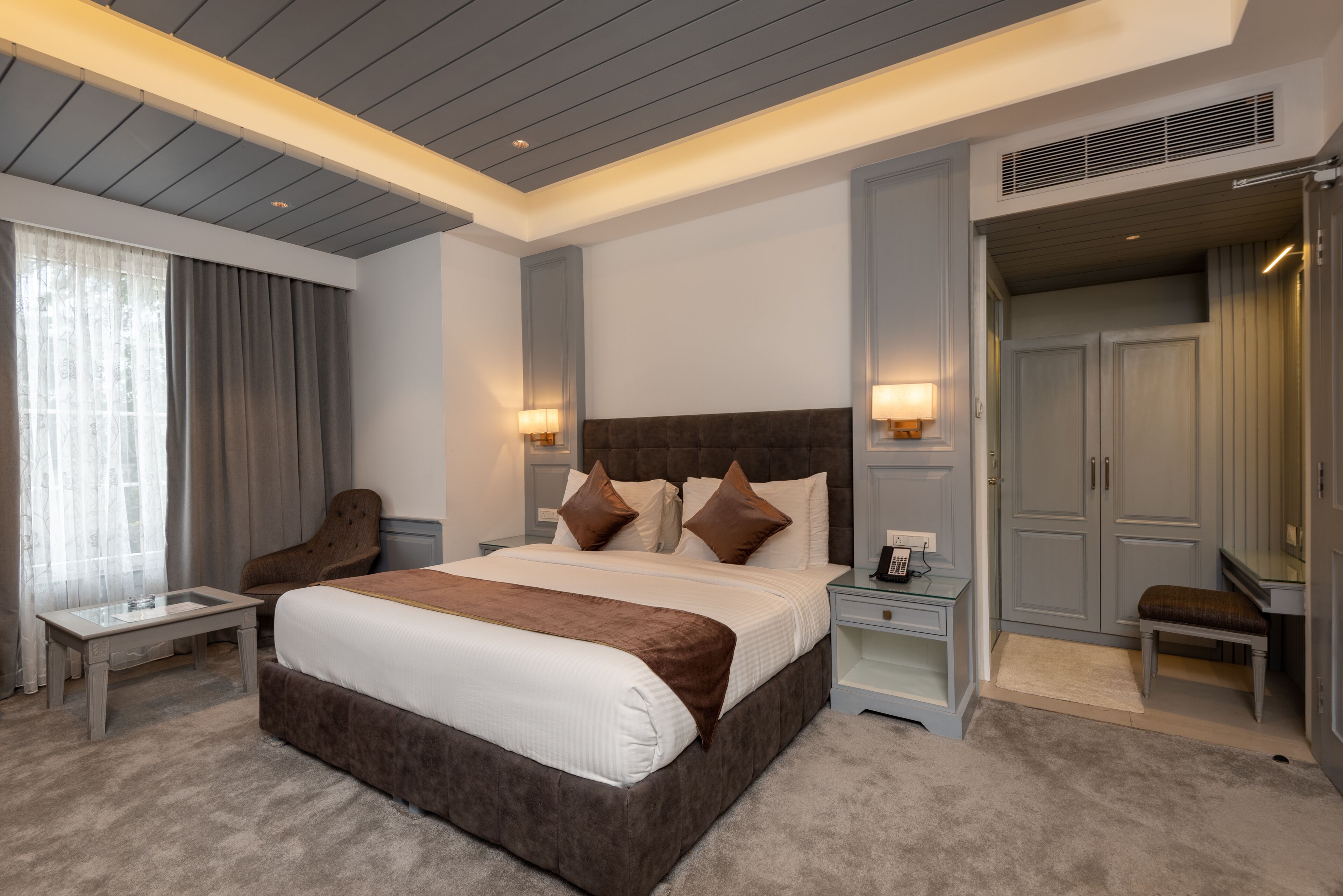 A modern hotel room with a large bed, bedside tables, a chair, a desk, and a wardrobe.