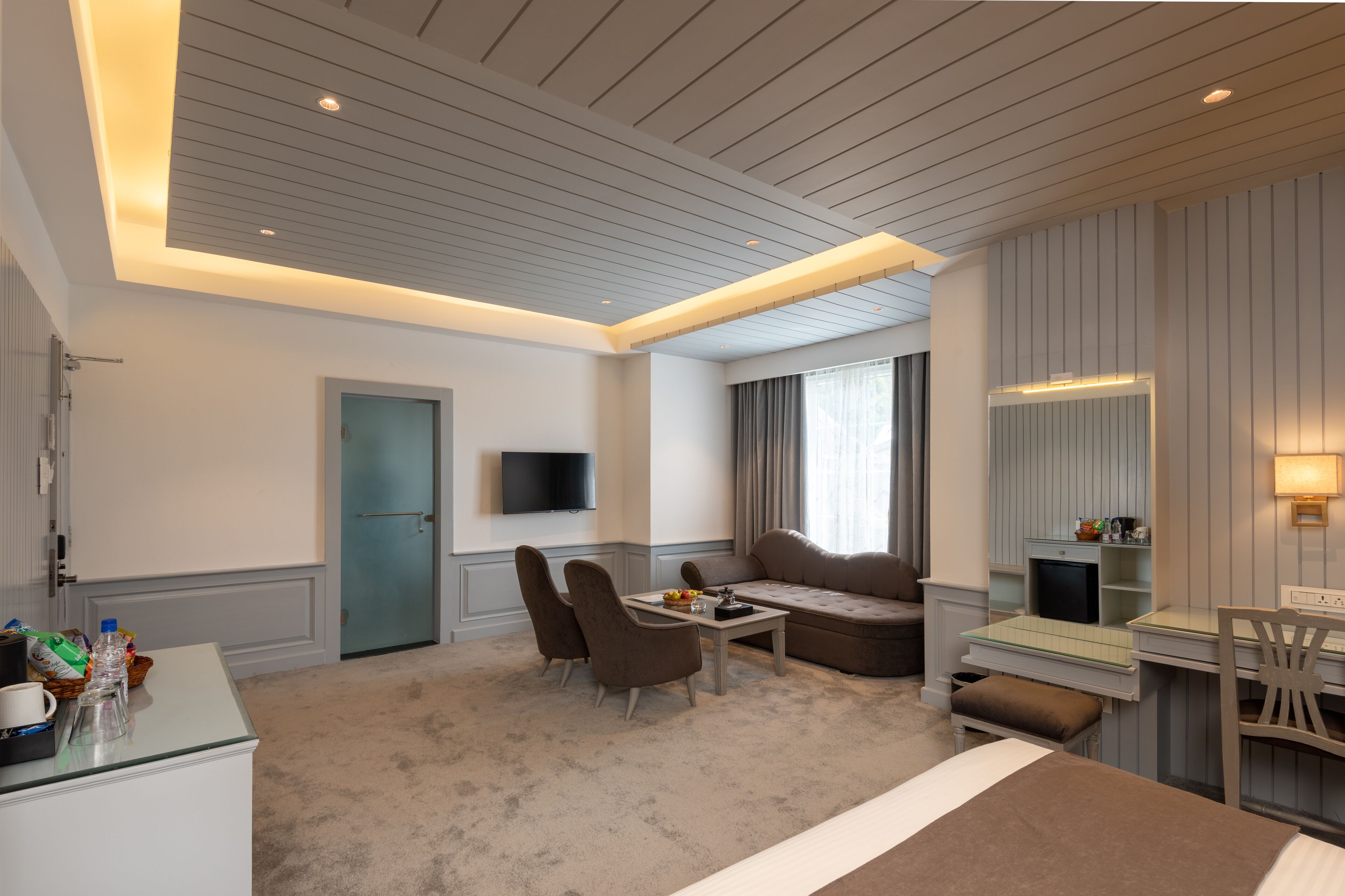 A modern hotel room with a sofa, a dining table, a TV, and a desk with a chair.