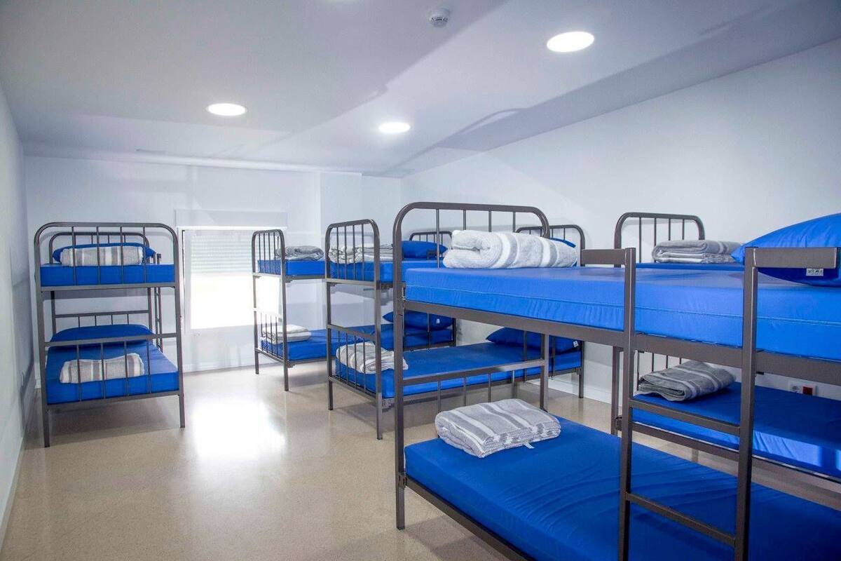 A dormitory room with bunk beds, each with a blue mattress and pillows, and a white blanket folded on the lower bunk.