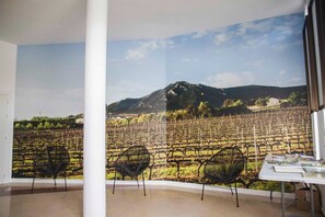A room with a large window overlooking a vineyard and a mountain. There are three black chairs and a table with books. - Albergue de Peregrinos San Cipriano - Hostel (Ayegui)