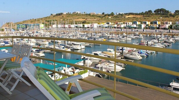Un puerto deportivo con numerosas embarcaciones amarradas, un balcón con sillones de descanso y vistas a edificios en una colina al fondo.