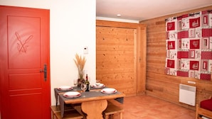 A dining area with a wooden table set for four, a red door with a floral design, and a wooden wall with a red and white patterned curtain. - Ground floor, balcony, parking, tv, ski locker, 39m², Pra Loup (Uvernet-Fours)