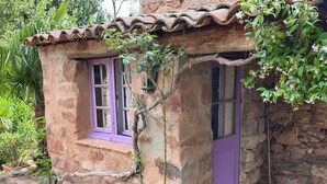 A rustic stone building with a tiled roof, a window with purple shutters, and a door with a broom leaning against it. - Two little romantic cottages in the middle of nature  (Les Mayons)