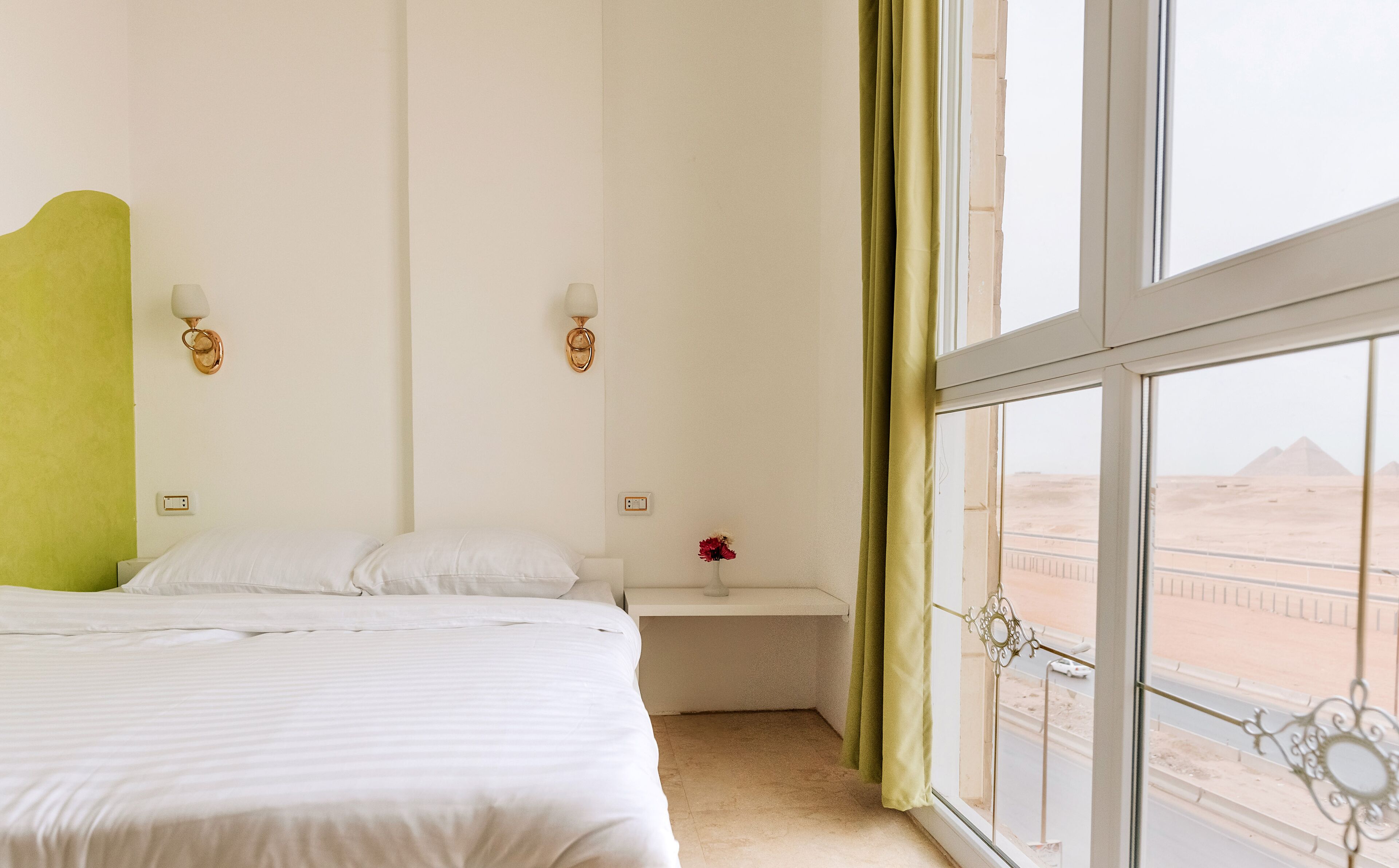 Ein Hotelzimmer mit großem Fenster, einem Bett, einem Nachttisch und Blick auf die Wüste.