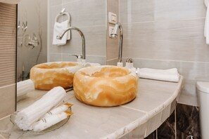 A bathroom with two large, round, beige-colored bathmats on the counter, a glass bowl with white towels, and a mirror reflecting the bathroom fixtures.