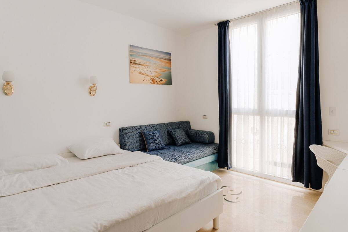 Ein schlicht eingerichtetes Schlafzimmer mit Bett, Sofa, Schreibtisch und einem großen Fenster mit Vorhängen.